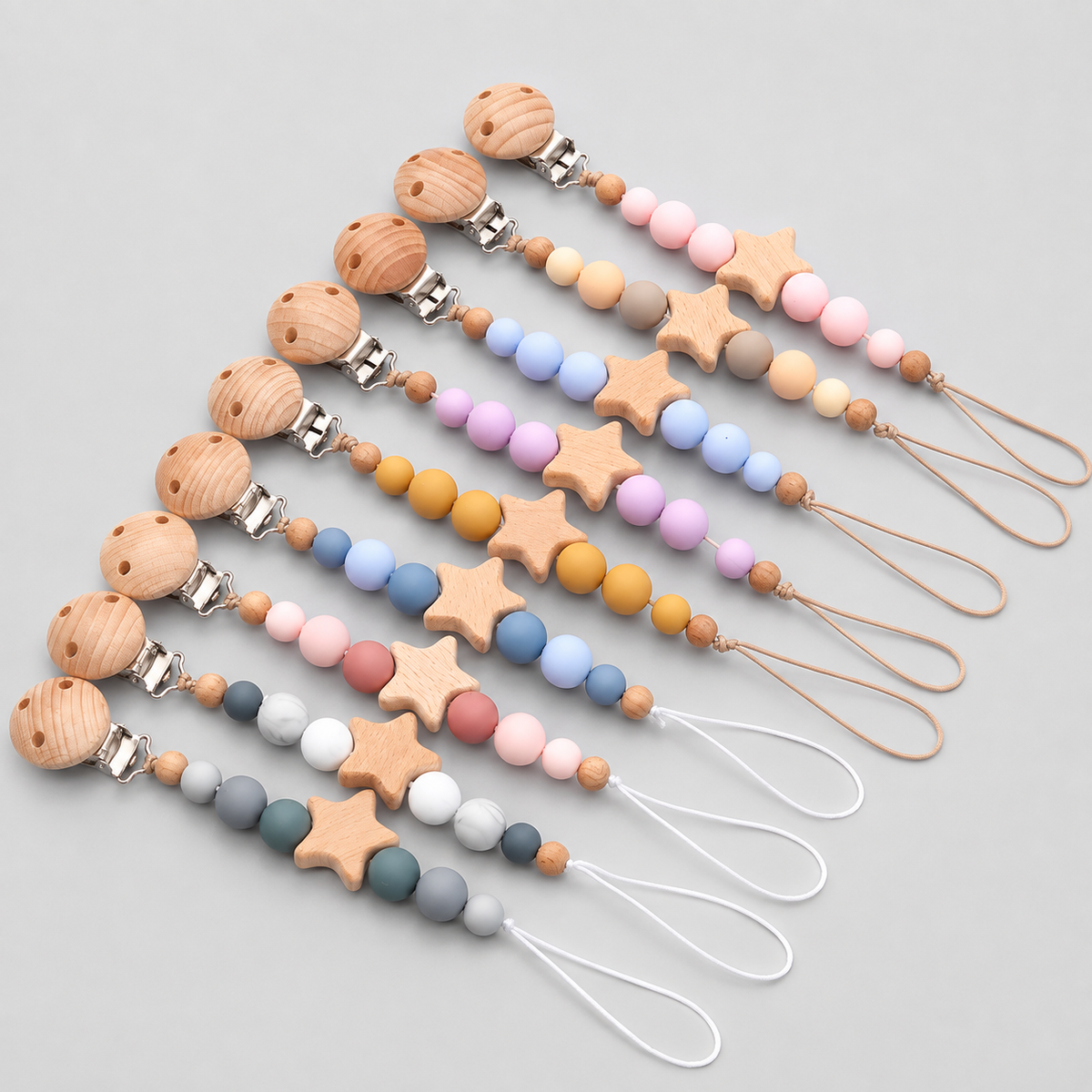 Six wooden baby teething keychains arranged diagonally on a light gray background. Each keychain features a round natural wood bead, a star-shaped wooden bead, and smooth pastel-colored silicone beads in various colors, finished with silver metal clasps and white or tan loops.